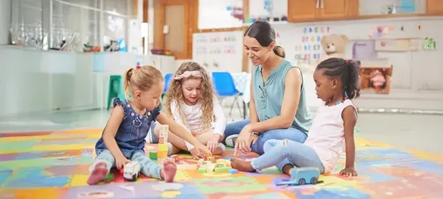 Preschool teacher interacting with a group of young children playing with building blocks on a colorful mat in a classroom setting.